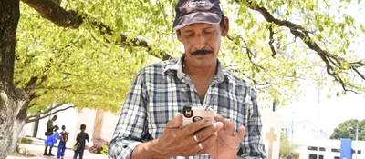 Hombre rural usa un celular bajo un árbol, mientras al fondo caminan personas en espacio público de un municipio