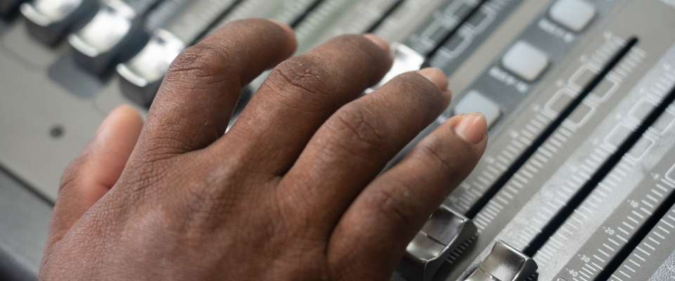 Primer plano de una mano operando una consola de audio. La imagen representa la radiodifusión comunitaria y la comunicación local.