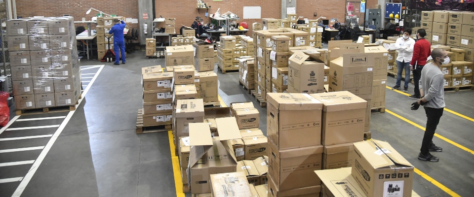 Bodega logística con decenas de cajas apiladas y trabajadores organizando envíos en amplios pasillos señalizados