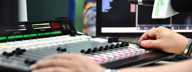 Persona opera una consola de audio en una cabina de radio, ajustando controles frente a monitores y un micrófono profesional.