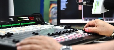 Persona opera una consola de audio en una cabina de radio, ajustando controles frente a monitores y un micrófono profesional.
