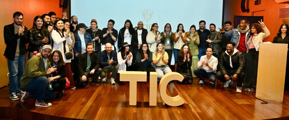 Grupo de personas posa y aplaude en un auditorio junto a las letras TIC, durante el anuncio de proyectos audiovisuales para TV pública.