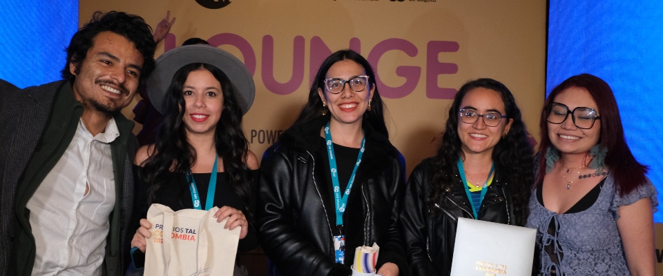 Cinco asistentes sonríen frente al panel de un evento, con acreditaciones y materiales, tras anuncio de nominados a Premios TAL.
