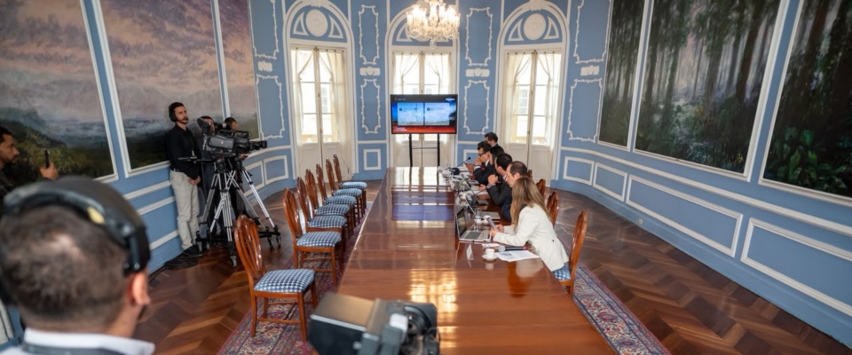 Sala de reuniones con mesa larga, funcionarios y periodistas. Al fondo, una pantalla durante una rueda de prensa oficial.