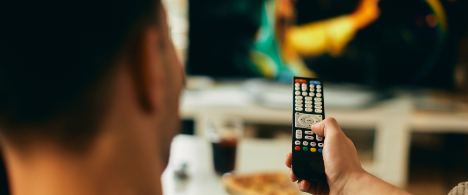 Persona de espaldas sostiene un control remoto frente a un televisor desenfocado. Al fondo hay un vaso y un plato sobre una mesa