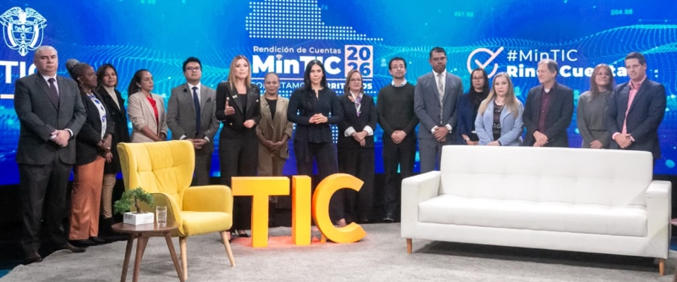 Catorce personas de pie detrás de sillón blanco, silla amarilla y letras TIC amarillas, frente a pantalla azul con textos