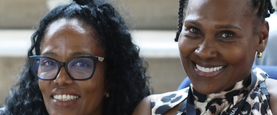 Dos personas sonríen a la cámara; izquierda con gafas negras, derecha con blusa estampada y arete dorado; fondo desenfocado.