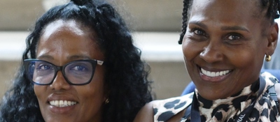 Dos personas sonríen a la cámara; izquierda con gafas negras, derecha con blusa estampada y arete dorado; fondo desenfocado.