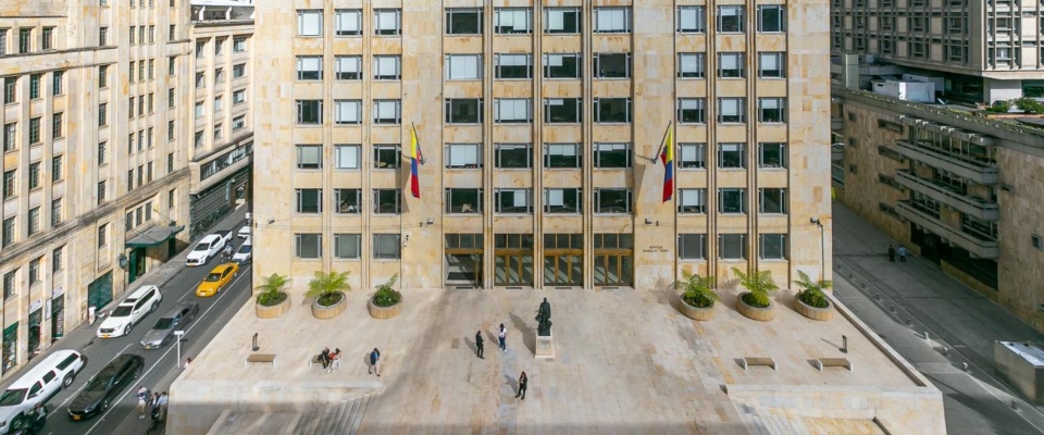 Edificio sede del Ministerio TIC con dos banderas, plaza de piedra, jardineras, estatua central, varias personas caminando y vehículos en calles laterales.