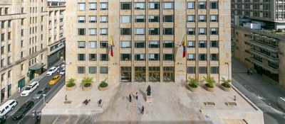 Edificio sede del Ministerio TIC con dos banderas, plaza de piedra, jardineras, estatua central, varias personas caminando y vehículos en calles laterales.