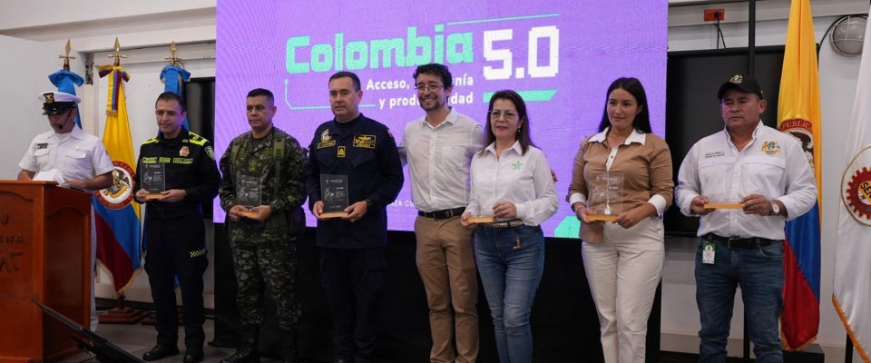 Nueve personas posan frente a pantalla morada "Colombia 5.0"; seis sostienen reconocimientos. Banderas y atril a los lados.