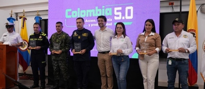 Nueve personas posan frente a pantalla morada "Colombia 5.0"; seis sostienen reconocimientos. Banderas y atril a los lados.