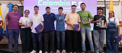 Nueve personas posan en tarima con placas y carpetas; al fondo, pantalla “Colombia 5.0” y banderas a ambos lados