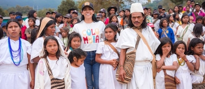 Grupo de personas con ropa blanca; mujer con camiseta "COLOMBIA" al centro. Niñas y niños alrededor, multitud al fondo.