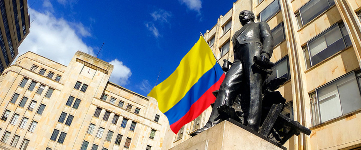 Estatua de bronce frente a edificios de piedra, con bandera de Colombia al centro y cielo azul con nubes.