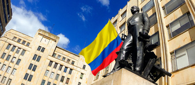 Estatua de bronce frente a edificios de piedra, con bandera de Colombia al centro y cielo azul con nubes.