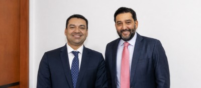Dos hombres posan de pie y sonríen, con vestimenta formal, frente a una bandera de Colombia