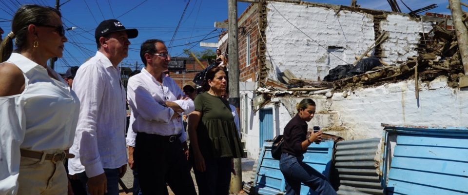 Cuatro personas observan una vivienda parcialmente derrumbada; otra mujer está sentada frente a la fachada dañada