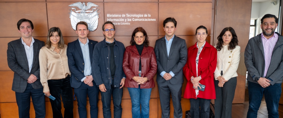 Nueve personas posan de pie frente al logo y nombre del Ministerio TIC; visten prendas en tonos oscuros, rojo, beige y gris