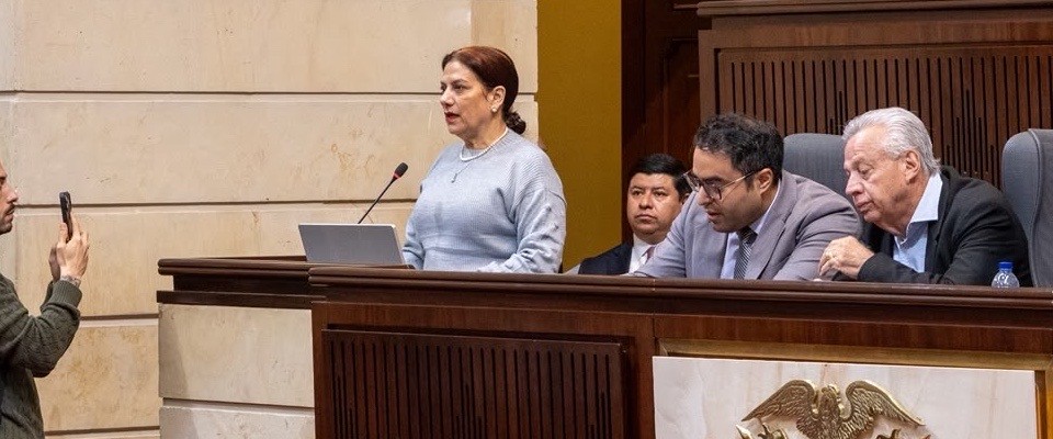 Mujer habla ante un micrófono con computador; cuatro hombres están cerca, uno sostiene un celular. Mobiliario de madera.