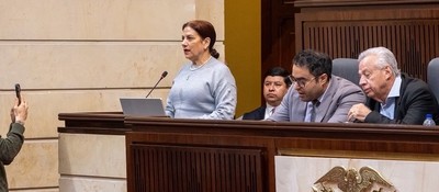 Mujer habla ante un micrófono con computador; cuatro hombres están cerca, uno sostiene un celular. Mobiliario de madera.