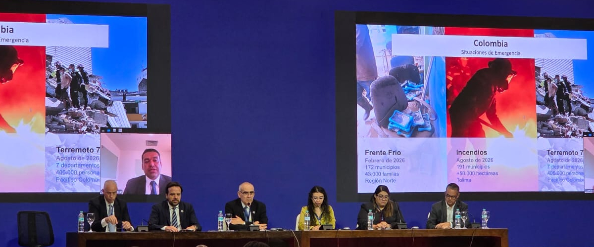 Seis personas están sentadas ante una mesa; detrás, dos pantallas muestran imágenes y datos sobre emergencias en Colombia.