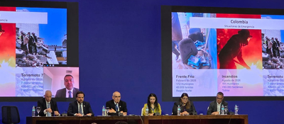 Seis personas están sentadas ante una mesa; detrás, dos pantallas muestran imágenes y datos sobre emergencias en Colombia.
