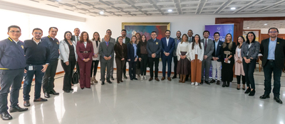Grupo de personas posa de pie en un salón; al fondo hay un cuadro, una pancarta y mobiliario.