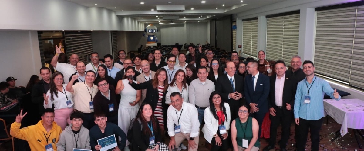 Grupo numeroso de personas posa para una foto en un salón; varias sonríen y algunas levantan las manos o muestran documentos.