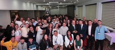 Grupo numeroso de personas posa para una foto en un salón; varias sonríen y algunas levantan las manos o muestran documentos.
