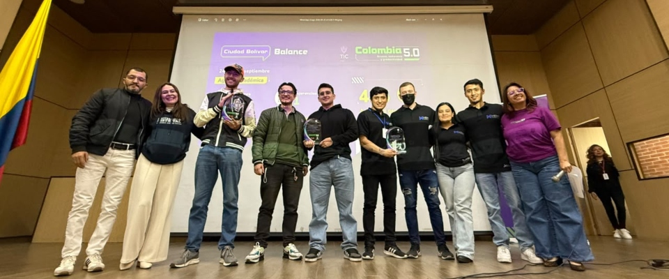 Nueve personas posan en un escenario frente a una pantalla con información de Colombia 5.0; algunas sostienen reconocimientos.