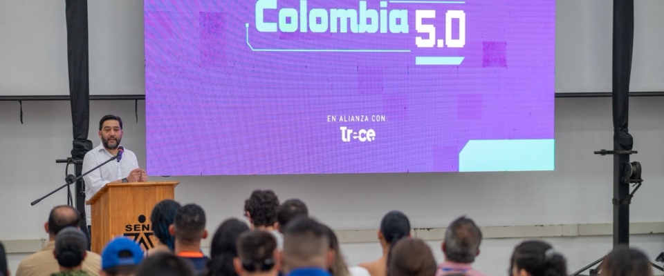 Hombre habla ante un público frente a una pantalla que muestra “Colombia 5.0” y “Trece”.
