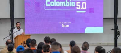 Hombre habla ante un público frente a una pantalla que muestra “Colombia 5.0” y “Trece”.