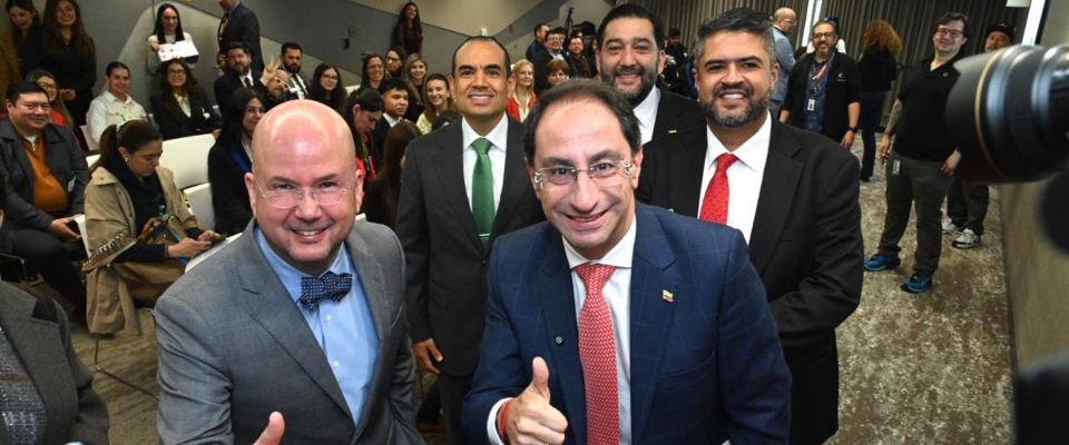 Cinco hombres con traje sonríen y dos hacen pulgar arriba; detrás hay asistentes sentados y de pie en una sala.