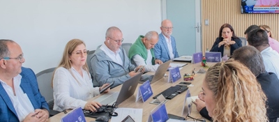 Varias personas reunidas alrededor de una mesa, con computadores portátiles, documentos y tarjetas, mientras algunas conversan.