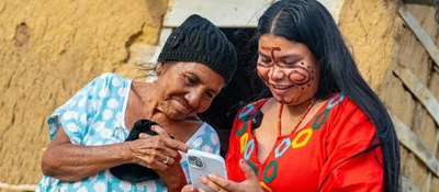 Mujer mayor y joven sonríen mientras miran un celular. Ambas tienen vestimenta colorida; la joven lleva pintura facial.