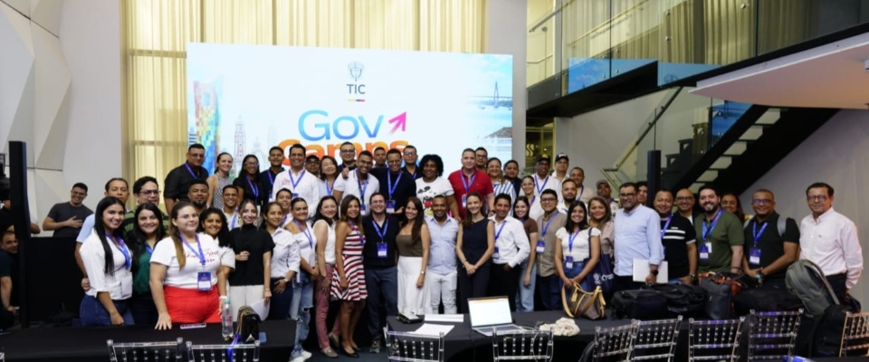 Fotografía grupal de participantes del evento GovCamp, realizado en un espacio institucional del sector TIC. Cerca de 50 personas posan frente a un pendón con la imagen de GovCamp.