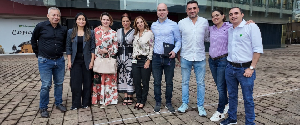 Ocho personas posan sonrientes frente a un edificio acristalado, sobre una plaza adoquinada; visten ropa formal e informal.