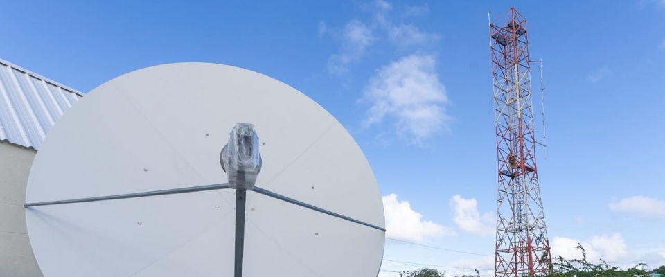 Antena parabólica blanca frente a una torre metálica roja y gris, junto a un edificio, bajo un cielo azul con nubes.